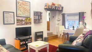 A living room with a fireplace, a television, a bookshelf, and a dining area with a table and chairs. - 1 bedroom accommodation in Stainforth near Settle (Stainforth near Settle)