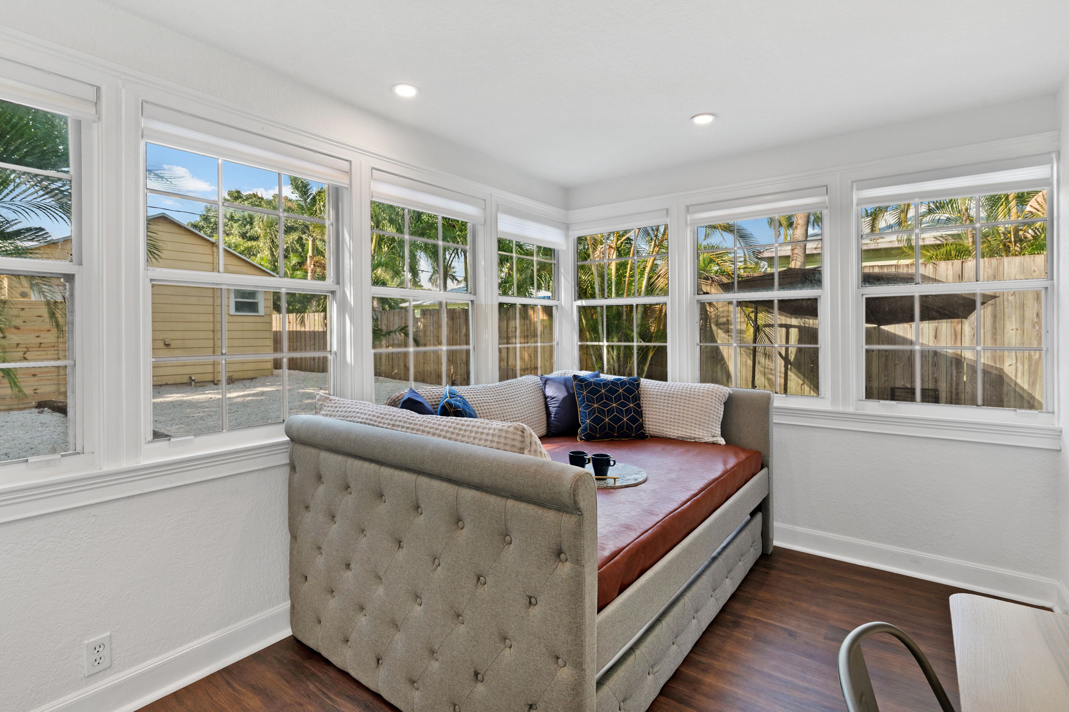 Una habitación con un banco empotrado y cajones, ventanas amplias con marcos blancos y vistas a un patio trasero con valla de madera y palmeras.