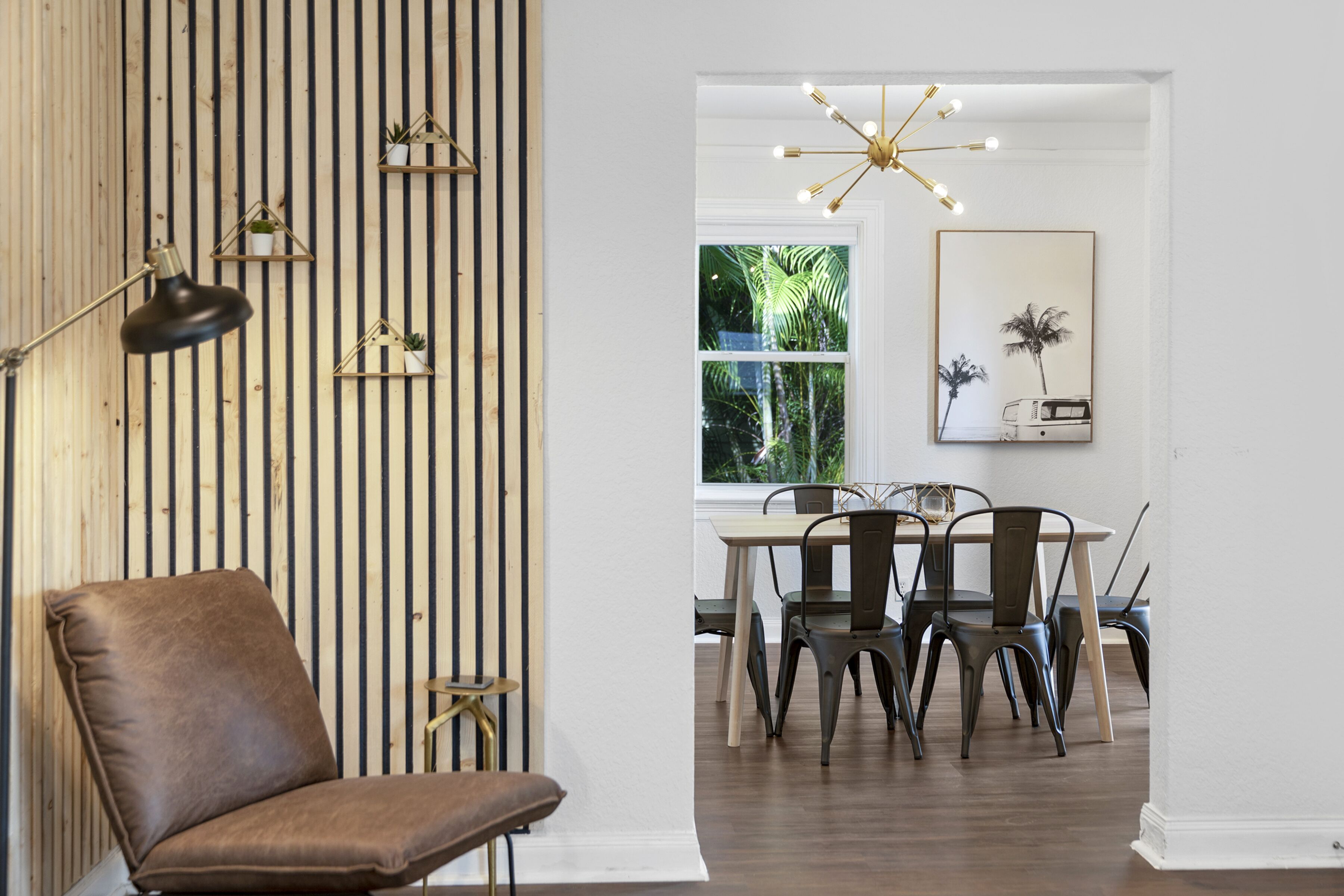 Un coin repas avec un plancher en bois, un fauteuil en cuir marron, une table avec des chaises en métal, un luminaire suspendu et une fenêtre donnant sur un paysage verdoyant.
