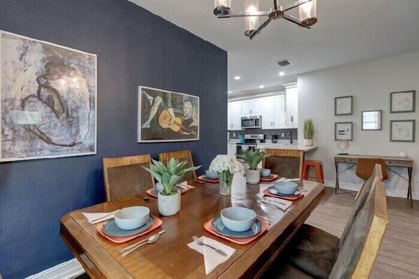 Une salle à manger avec une table en bois dressée pour six personnes, entourée de chaises. Deux tableaux ornent le mur, un ventilateur de plafond est suspendu au-dessus, et on aperçoit une cuisine au fond.
