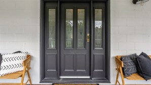 Una puerta principal gris oscuro con paneles de vidrio y un llamador de latón, flanqueada por dos bancos de madera con cojines estampados.