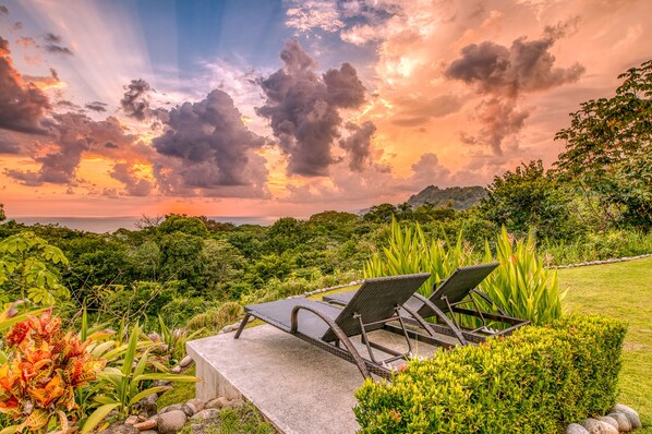 A garden with a concrete bench and two wicker chairs, surrounded by lush greenery and a view of the ocean at sunset.
