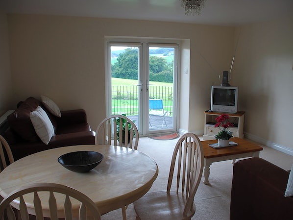 A living room with a sofa, a dining table, chairs, a TV on a stand, and a sliding glass door leading to a balcony.