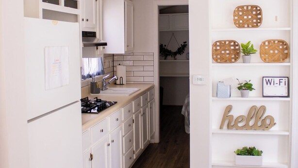 A kitchen with white cabinets, a stove, and a sink. There is a shelf with wicker baskets and a 'hello' sign. - Clyde's Cabin + Hot Tub (Carthage)