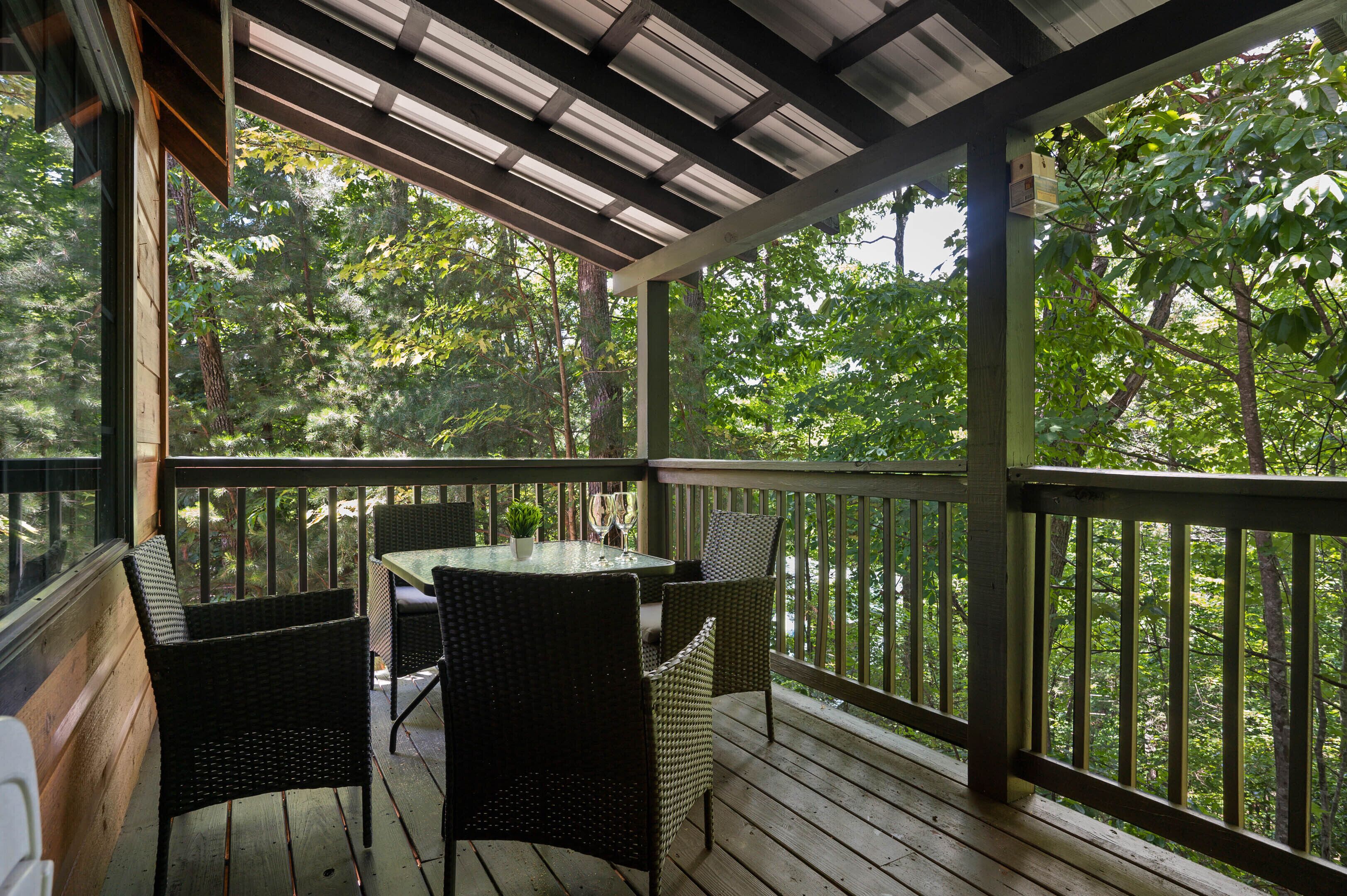 A covered outdoor area with a table and chairs, surrounded by wooden railings and overlooking a forested area.