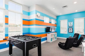A room with a foosball table, black chairs, a TV, and a window with blinds.