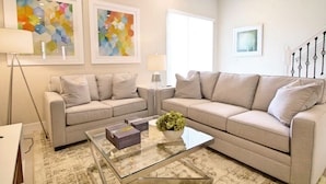 Ein modernes Wohnzimmer mit zwei beigen Sofas, einem Glastisch und abstrakter Kunst an den Wänden.