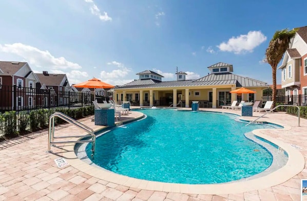 A swimming pool with lounge chairs and umbrellas, surrounded by a fence and houses.