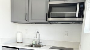 A modern kitchen with grey cabinets, a stainless steel refrigerator, a microwave, and a sink with a faucet. - Comfortable Tiny House in a Great Location (Moreno Valley)