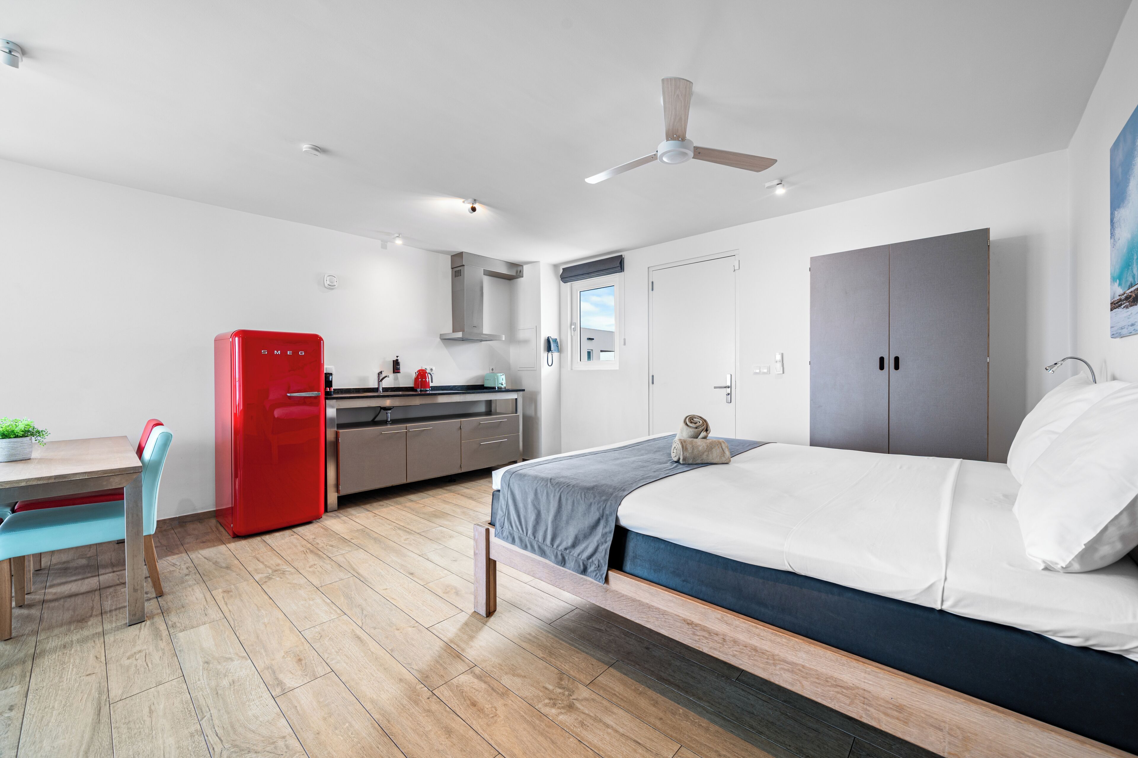 Une chambre moderne avec un lit, un bureau, un réfrigérateur rouge et un coin cuisine.
