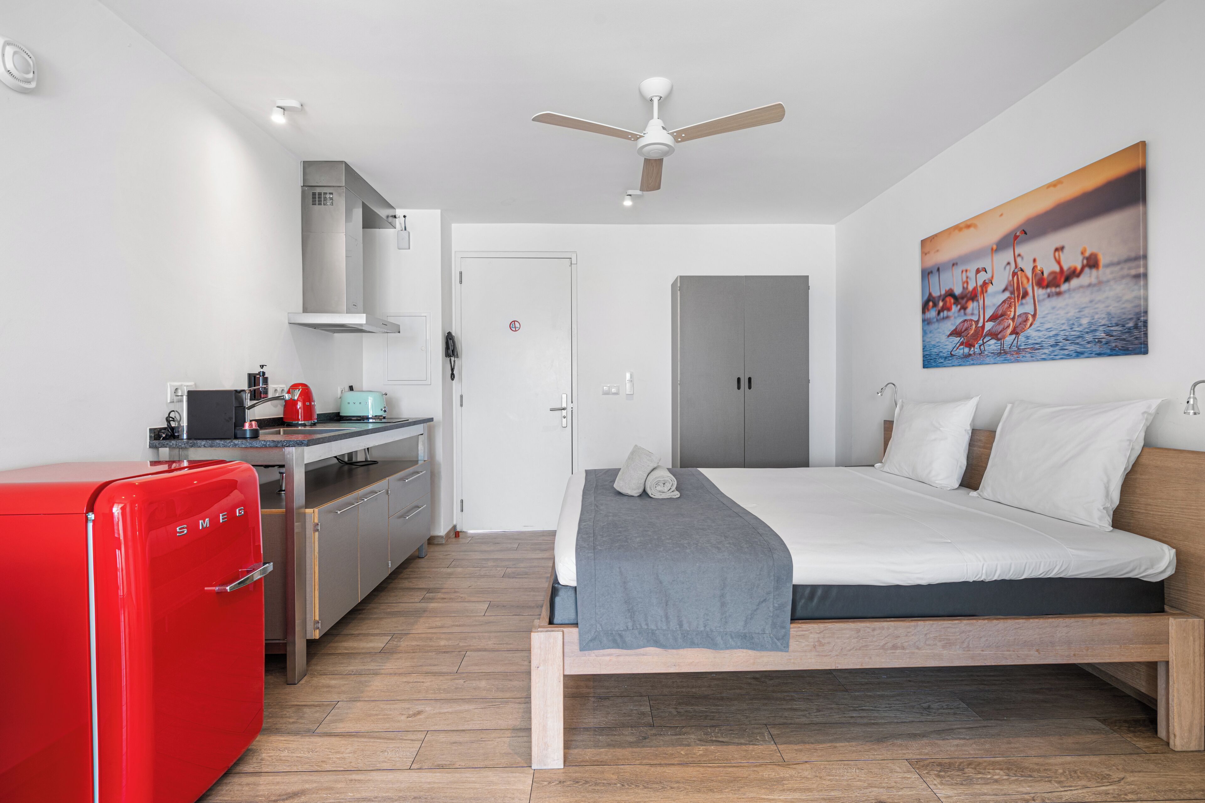 Quarto de hotel moderno com cama, frigorífico vermelho, cozinha compacta e ventoinha no teto.