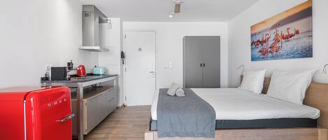 Quarto de hotel moderno com cama, frigorífico vermelho, cozinha compacta e ventoinha no teto.