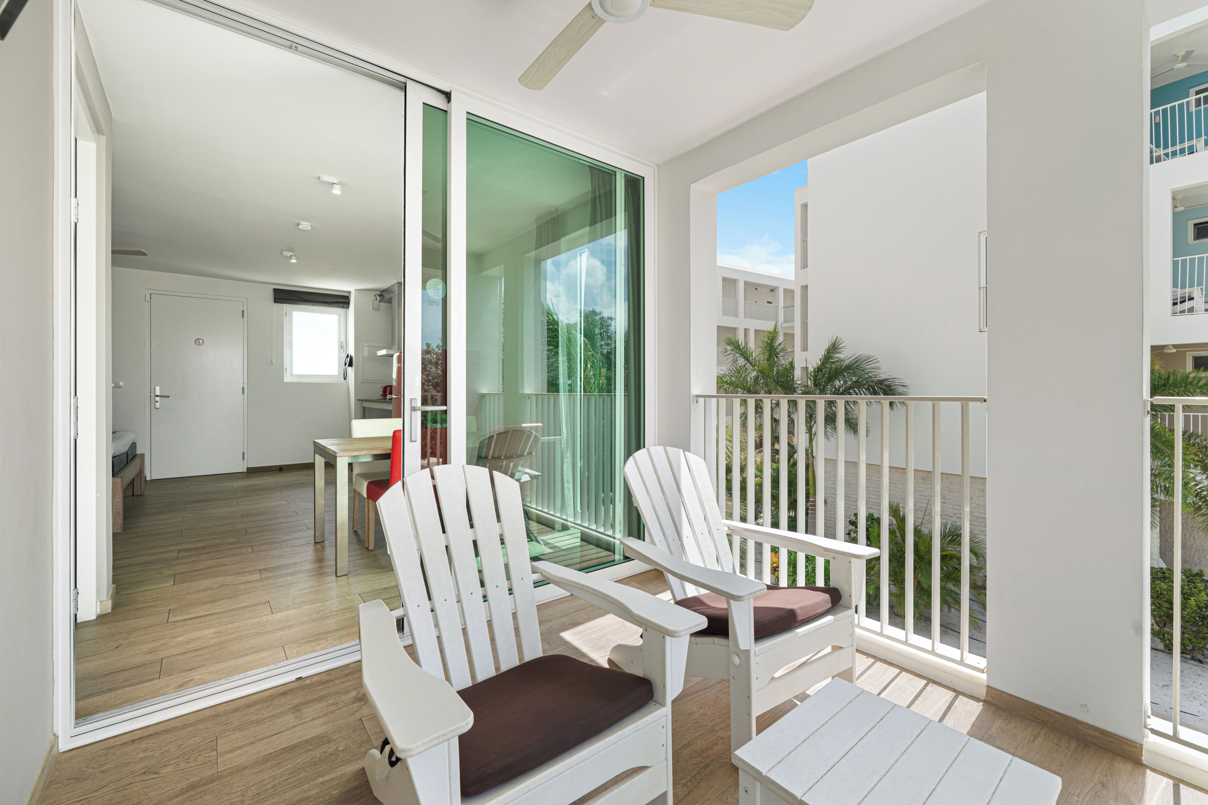 Een balkon met witte stoelen en een tafel, een glazen deur die naar een kamer leidt, en een uitzicht op een gebouw en groen.