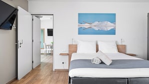 Una habitación de hotel moderna con piso de madera, una cama grande con cubrecama gris y blanco, un cuadro de montañas en la pared y un televisor montado en la pared.