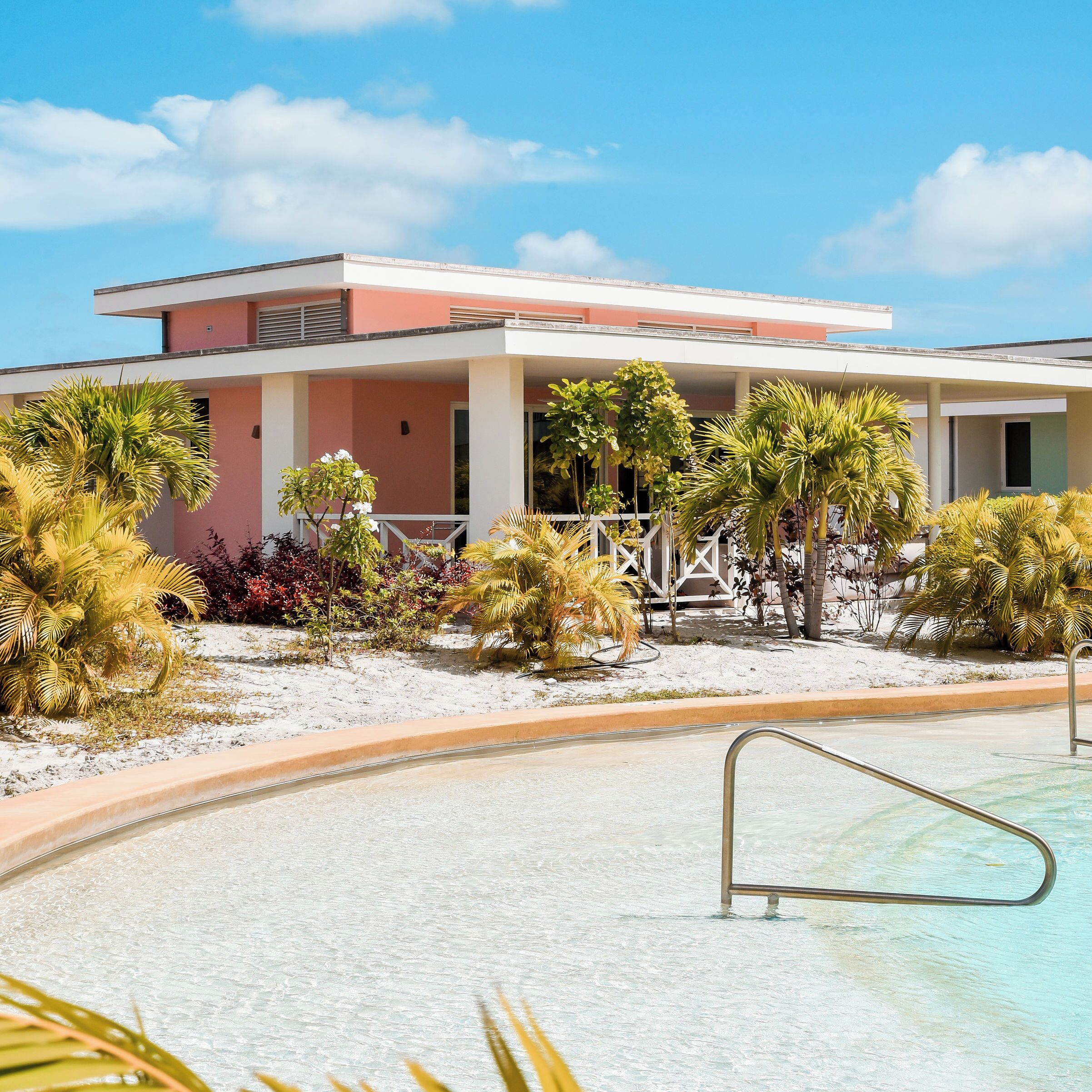 A modern hotel with a pool, surrounded by tropical plants and a clear sky.