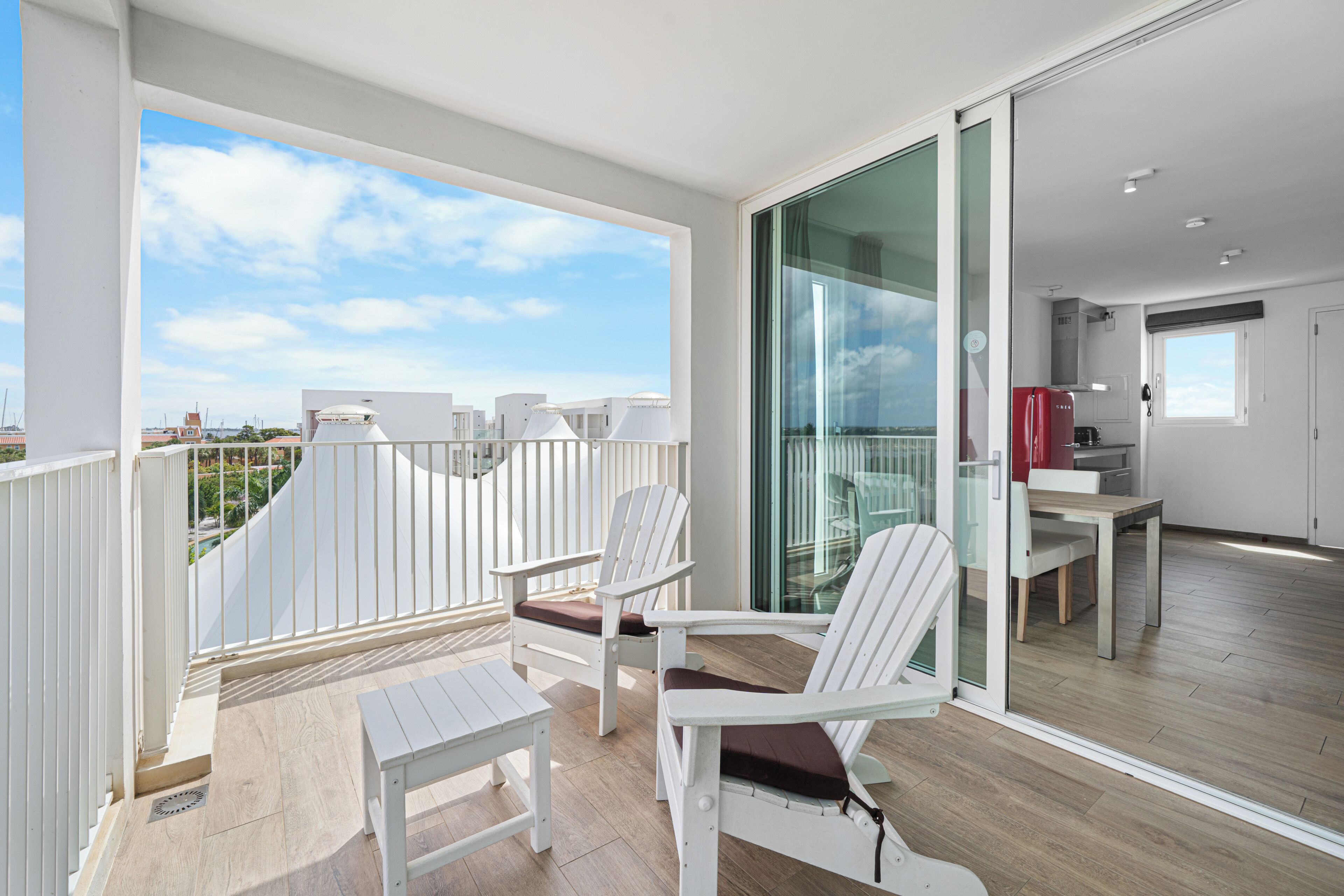 Ein Balkon mit weißen Stühlen und einem kleinen Tisch, eine Schiebetür, die zu einer Küche führt, und Blick auf die Stadtlandschaft.