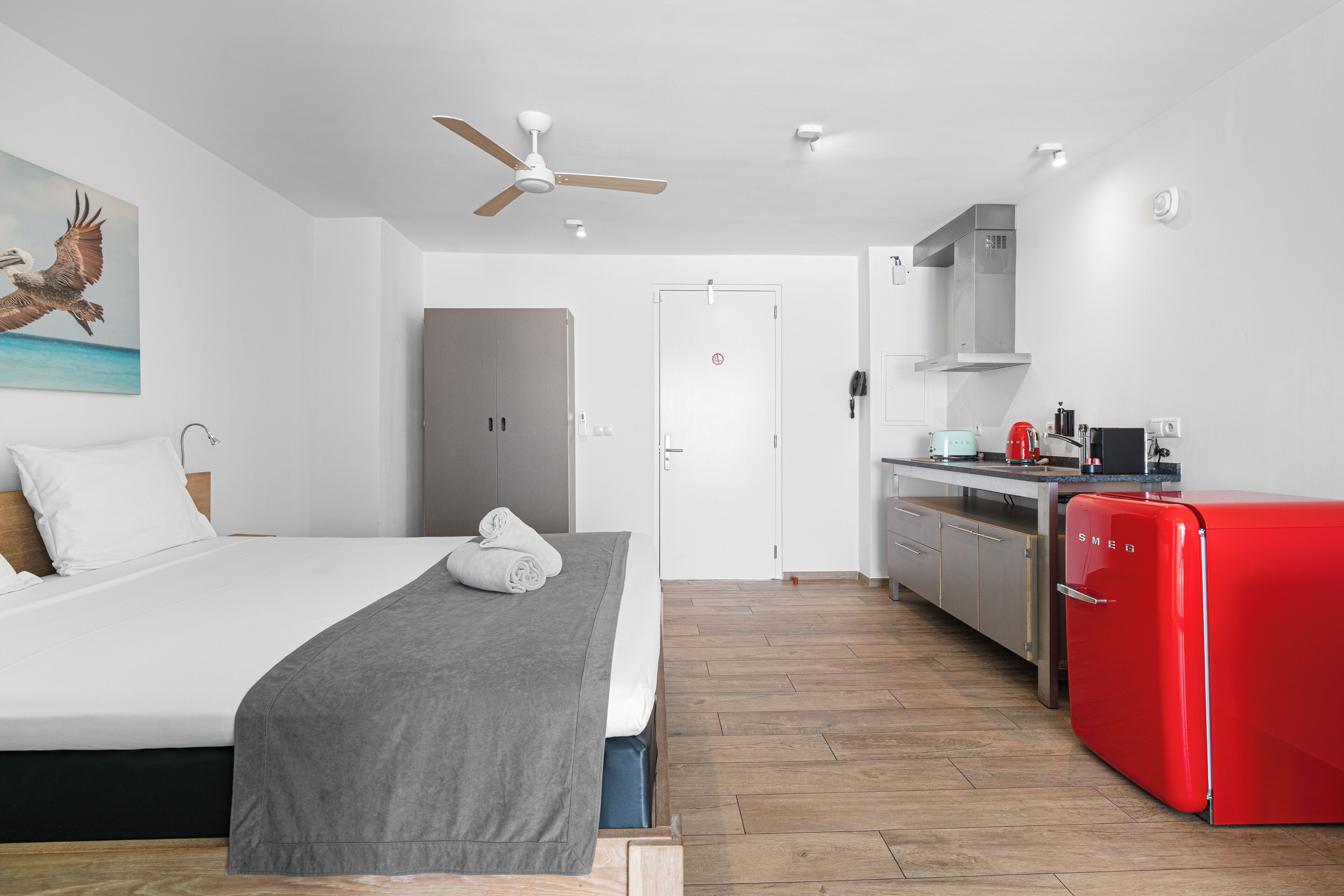A bedroom with a bed, a gray blanket, and a ceiling fan. Adjacent is a kitchen with a red refrigerator and a stove.