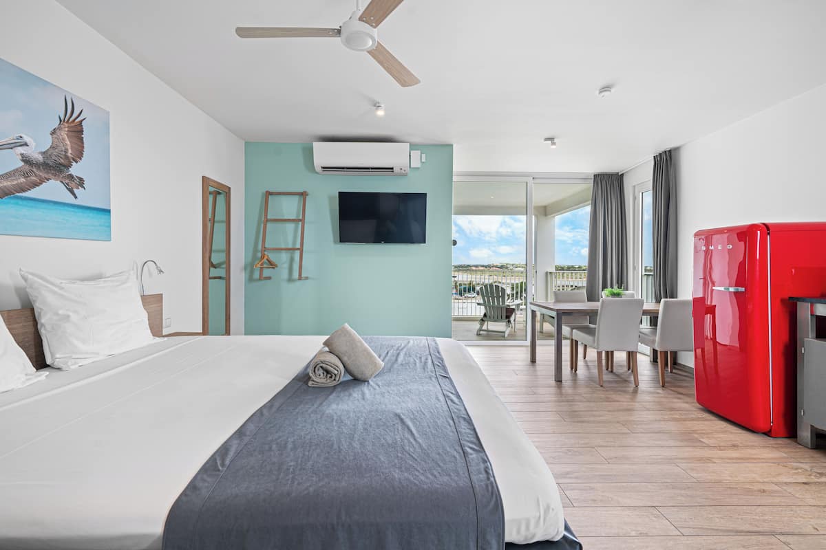 Une chambre d’hôtel avec un lit, une télévision, un ventilateur de plafond, un réfrigérateur rouge et un balcon équipé d’une table et de chaises.