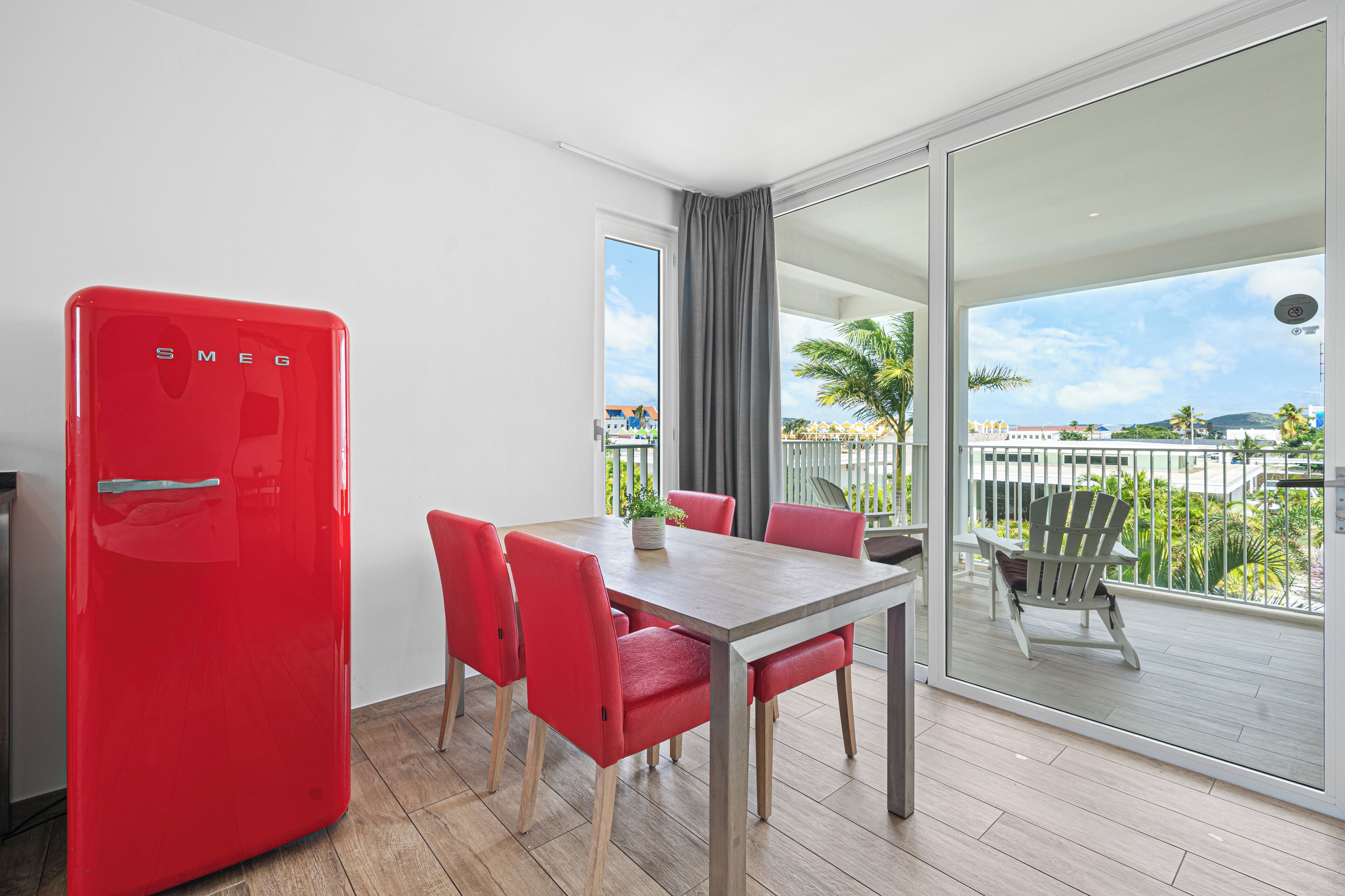 A modern room with a red refrigerator, a wooden dining table with red chairs, and a balcony with a view of the city and palm trees.