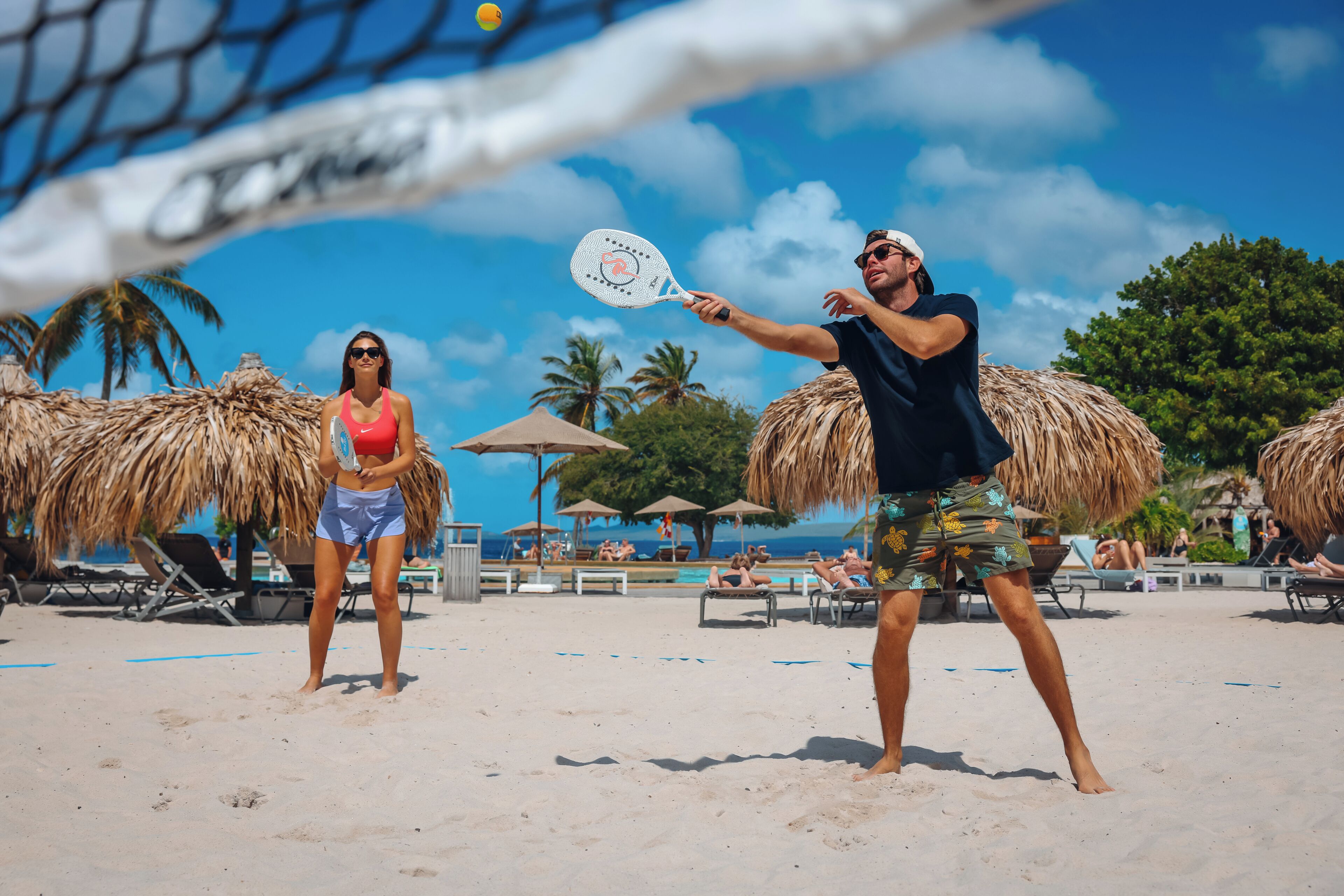 A beach volleyball game with a net, palm trees, and beach chairs in the background.