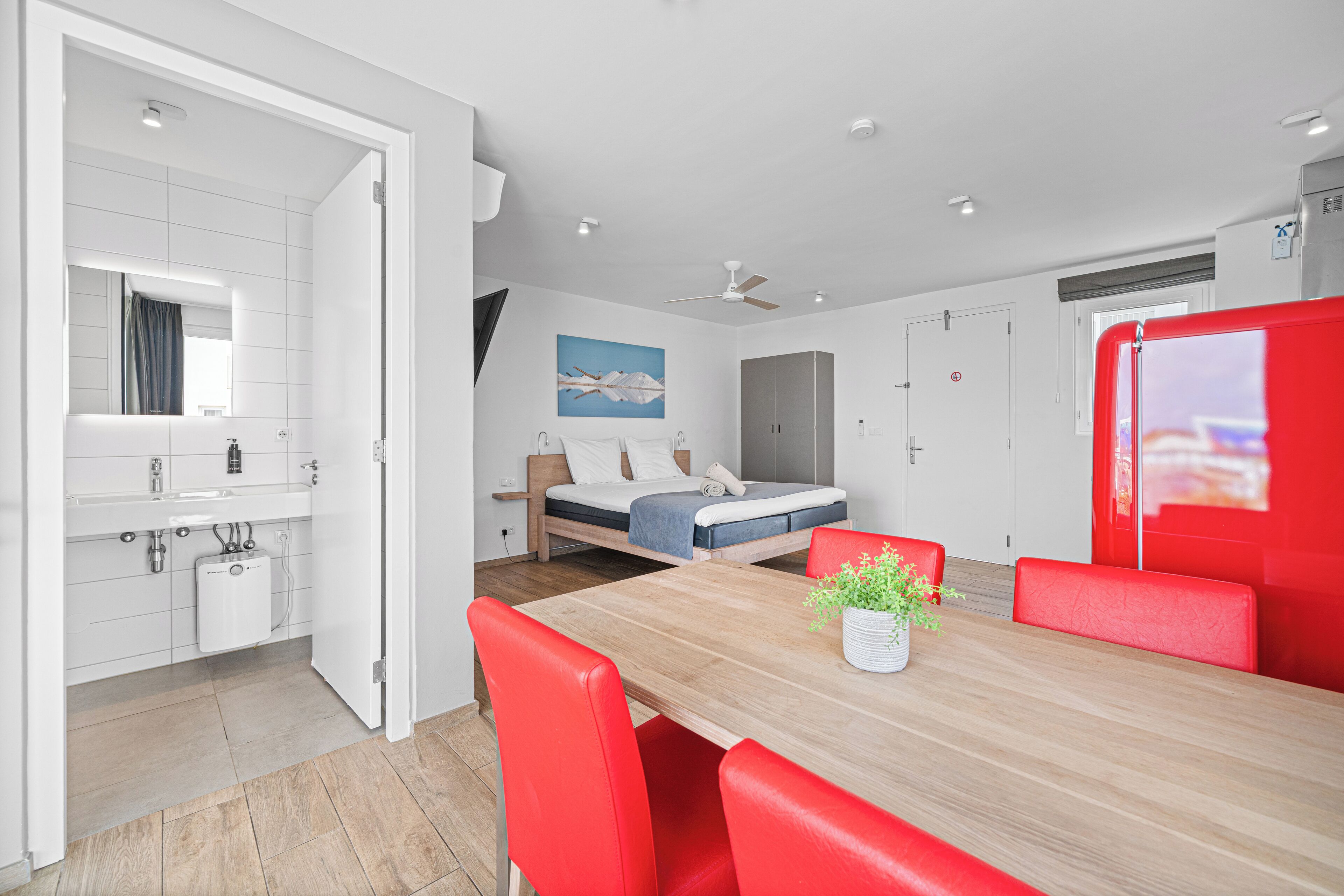 A modern hotel room with a wooden dining table, red chairs, a bed with a blue mattress, a white bathroom, and a ceiling fan.