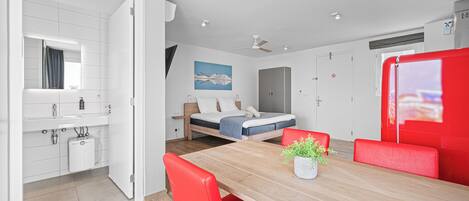 A modern hotel room with a wooden dining table, red chairs, a bed with a blue mattress, a white bathroom, and a ceiling fan.