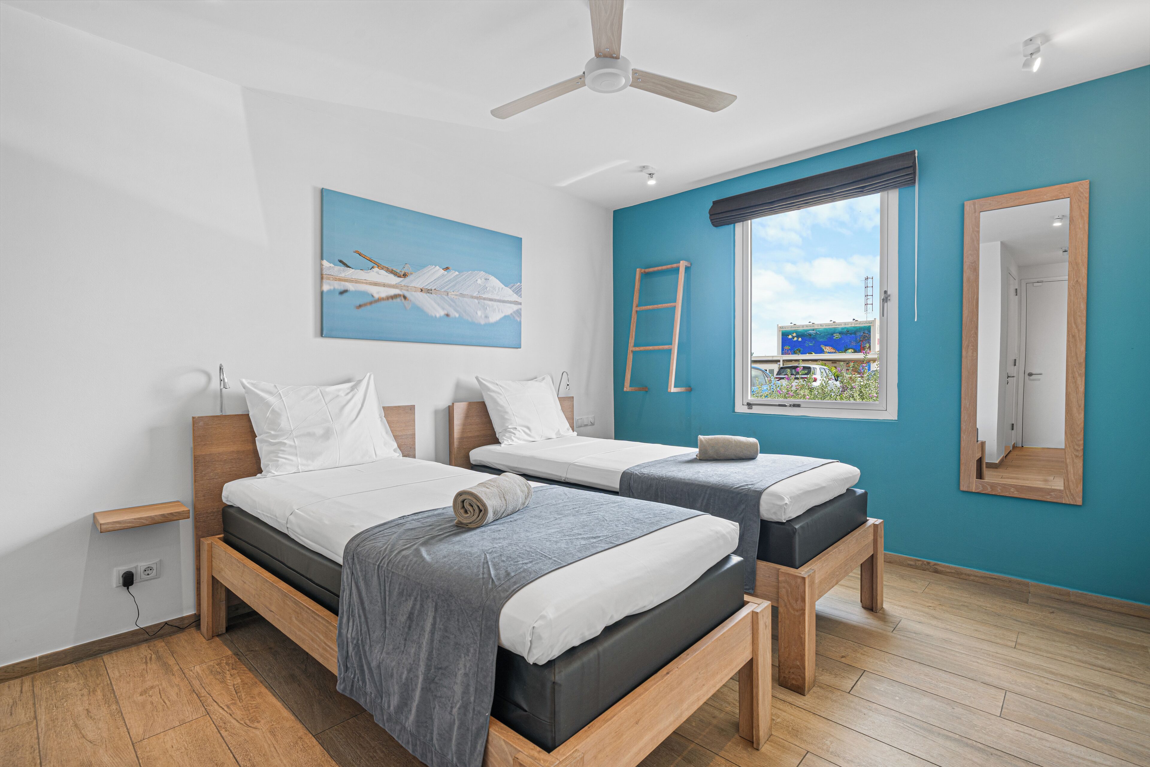 A modern bedroom with two beds, a ceiling fan, a window with a view, and a painting on the wall.