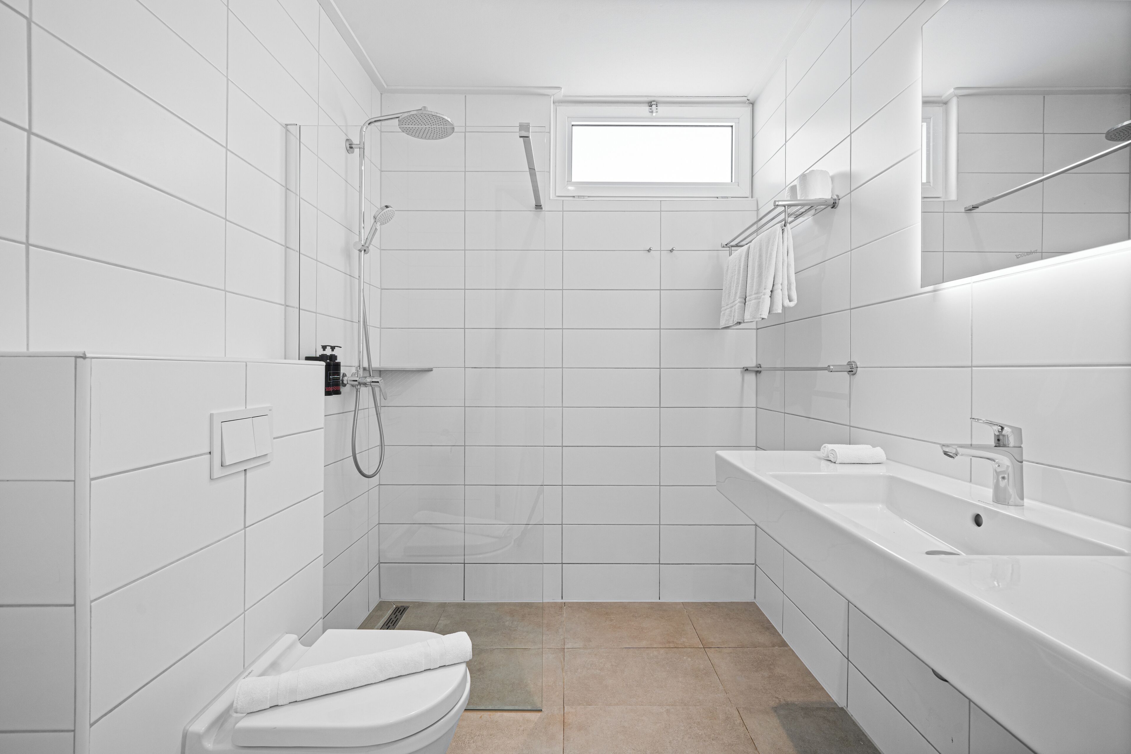 A modern bathroom with white tiles, a shower, a toilet, a sink, and a window.