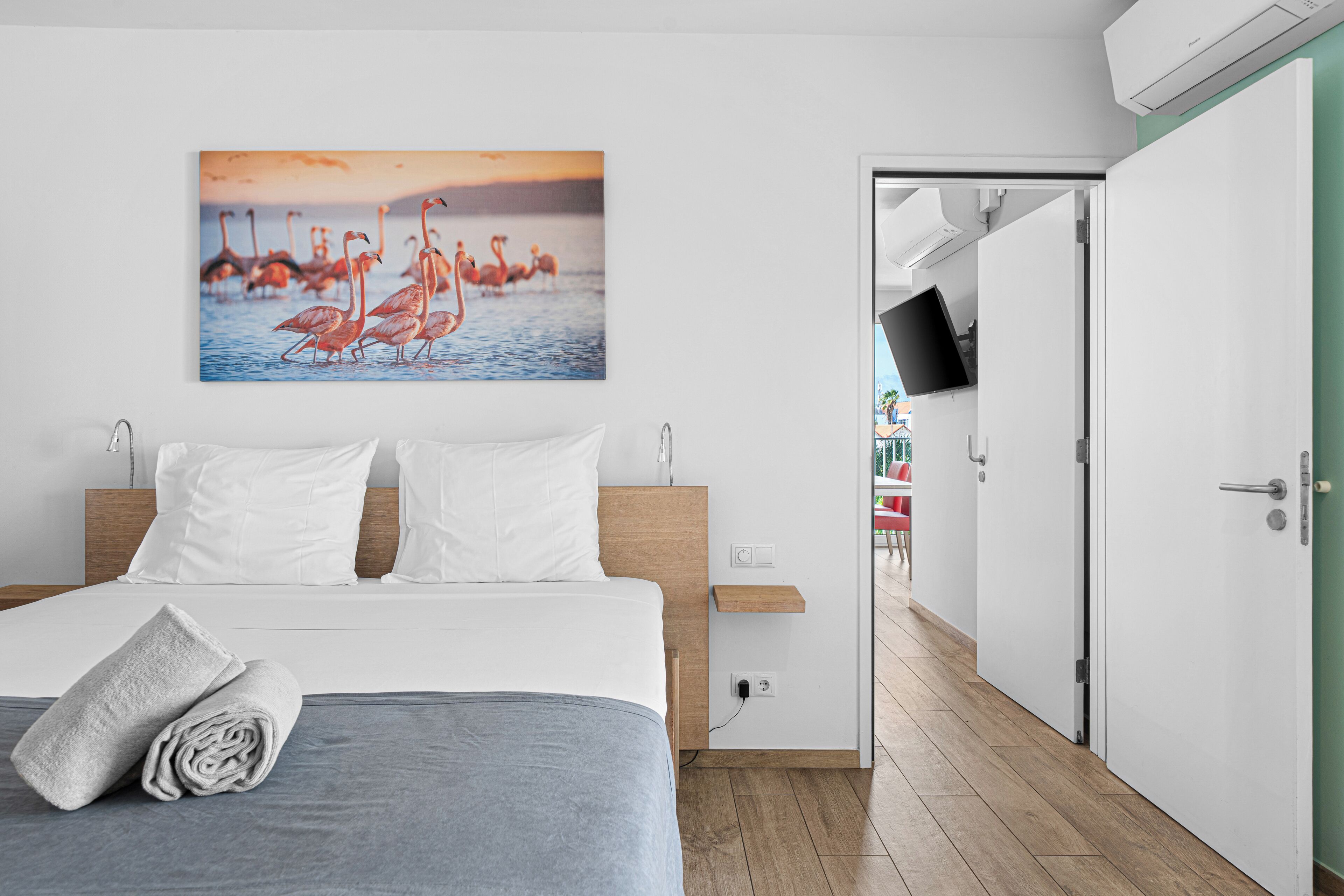 Une chambre d’hôtel avec un lit, des oreillers blancs, une couverture grise, une serviette pliée, un téléviseur fixé au mur et un tableau représentant des flamants roses.