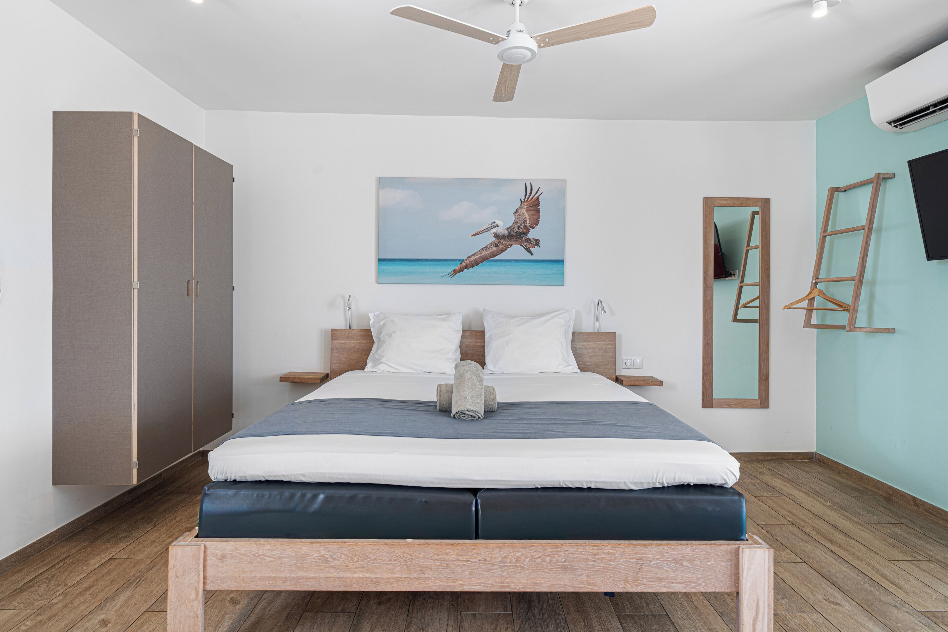 A bedroom with a bed, wooden wardrobe, a ceiling fan, a painting of a bird, a mirror, and a TV on the wall.