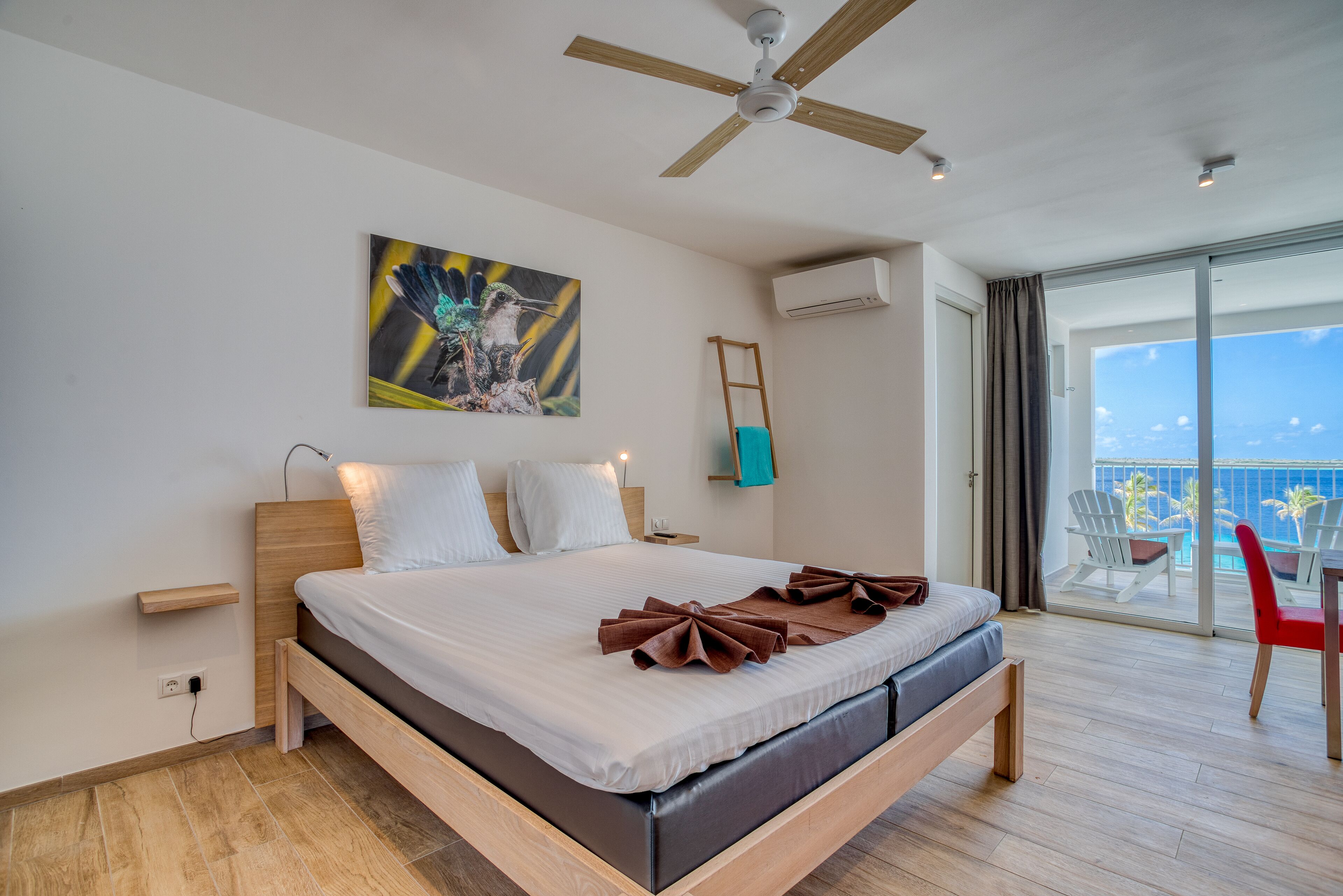 A modern hotel room with a large bed, a ceiling fan, a painting, an air conditioning unit, and a balcony with a view of the ocean.