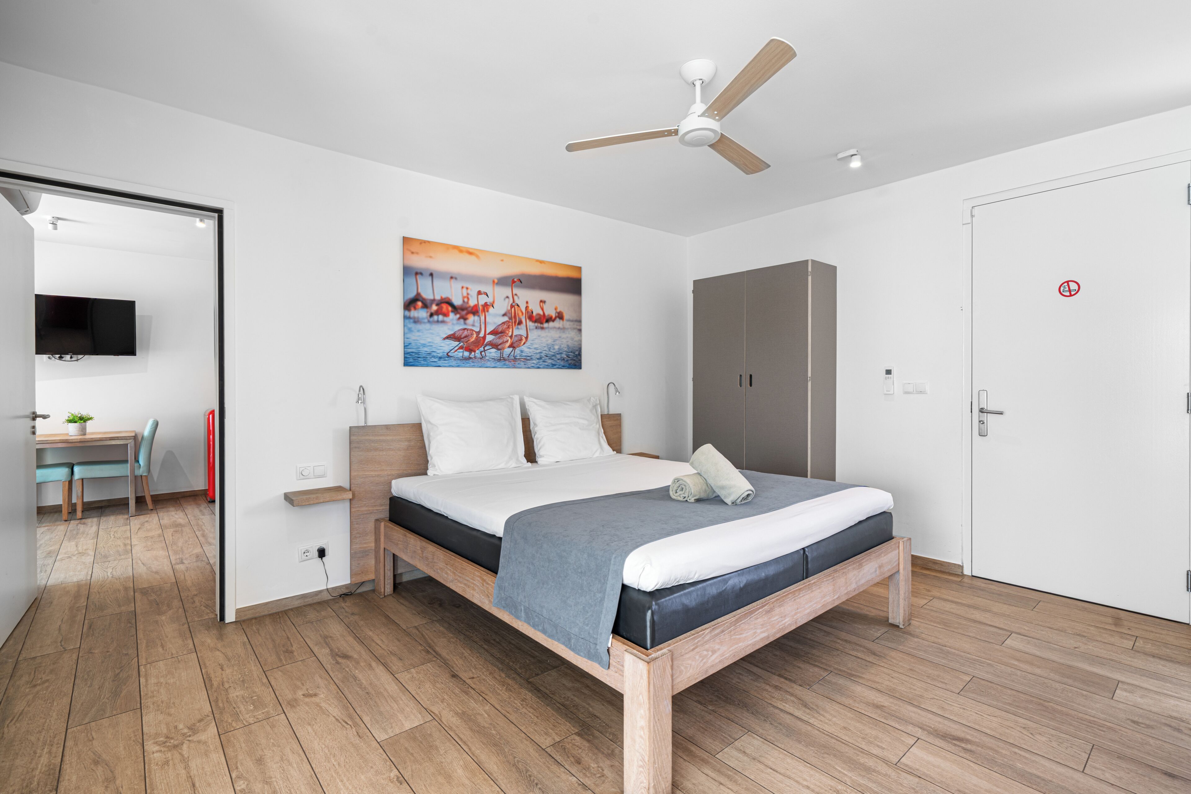 A modern bedroom with a wooden bed, a ceiling fan, a painting of flamingos, a wardrobe, and a desk with a chair.