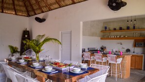 Un comedor con una larga mesa de madera dispuesta para seis personas, rodeada de sillas de mimbre blanco. Se ve una cocina con gabinetes de madera y una encimera al fondo.
