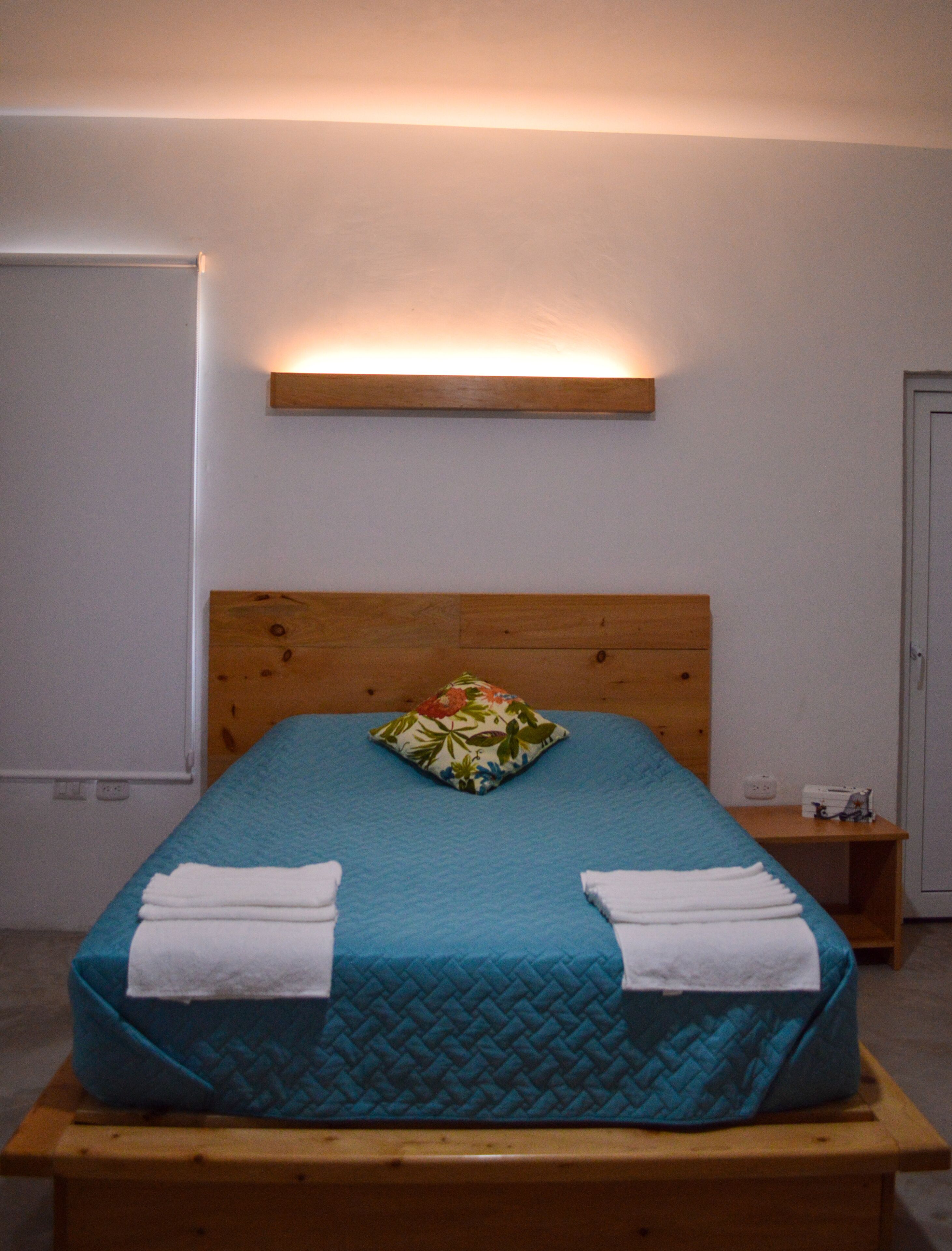 A bed with a wooden headboard, a teal bedspread, and a floral pillow. There are two white towels folded at the foot of the bed. A small wooden nightstand with a lamp is to the right of the bed.
