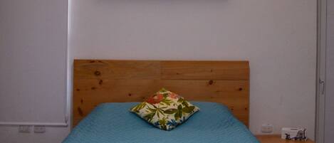 A bed with a wooden headboard, a teal bedspread, and a floral pillow. There are two white towels folded at the foot of the bed. A small wooden nightstand with a lamp is to the right of the bed.