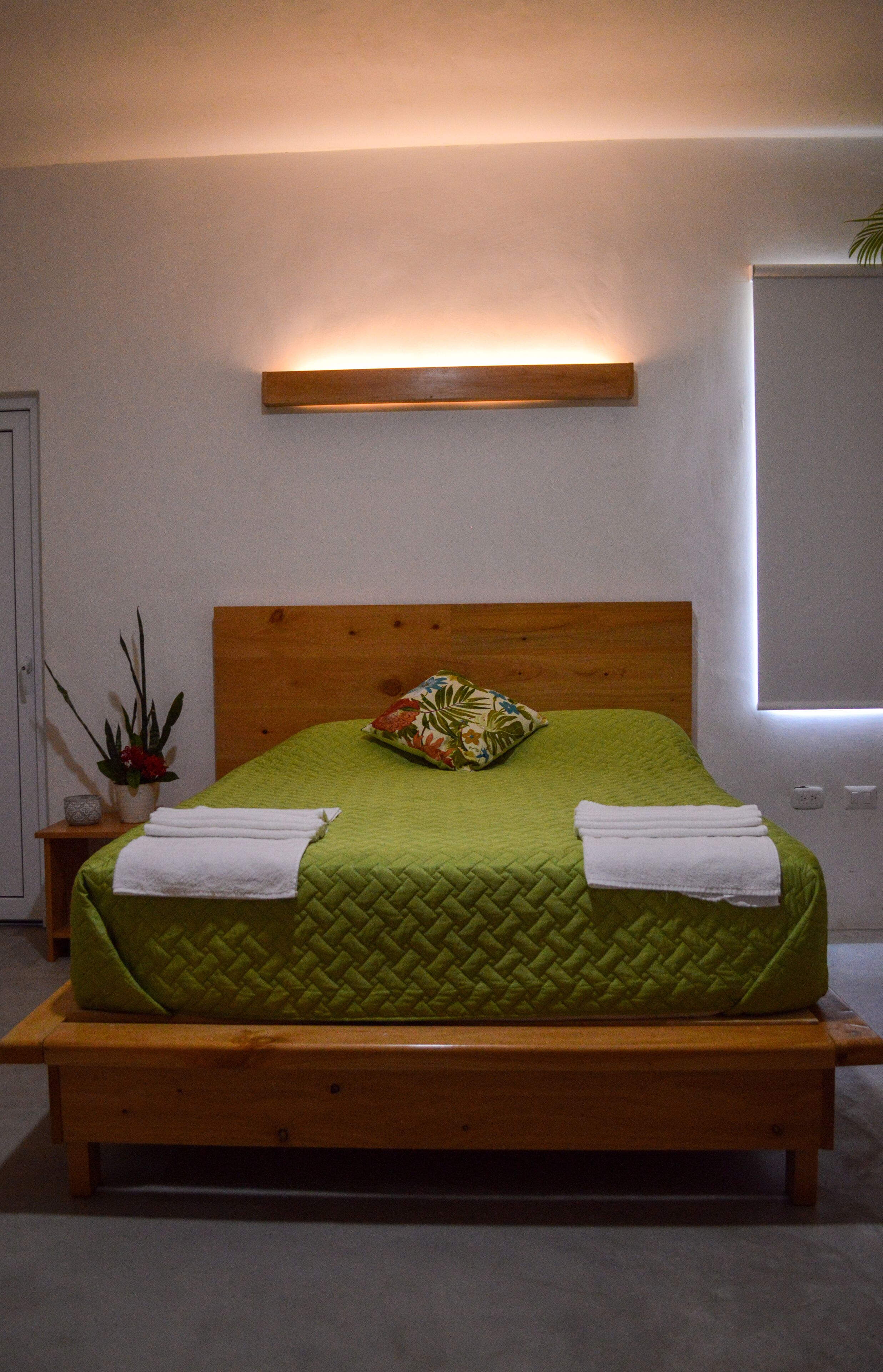 Un lit bien fait, avec une tête de lit en bois, une plante sur la table de chevet et un luminaire fixé au mur.