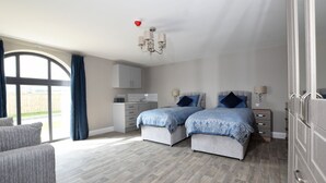 A bedroom with two beds, a sofa, a kitchen area, and a large window with curtains.