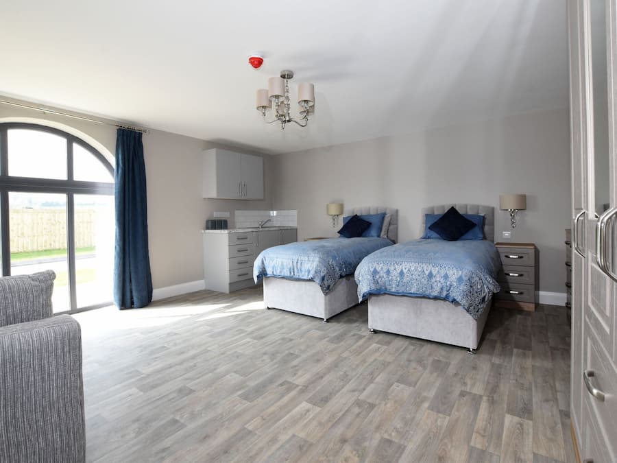 A bedroom with two beds, a sofa, a kitchen area, and a large window with curtains.