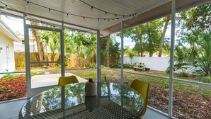 A covered patio with a round glass table and yellow chairs, overlooking a backyard with trees and a wooden fence. - ❤️ of Ormond Beach ⭐️Self check-in⭐️Beach (Ormond Beach)
