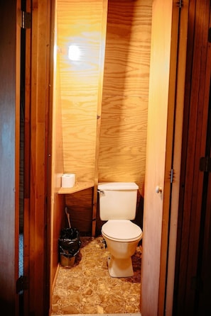A small, wooden-paneled bathroom with a toilet, a small shelf with toilet paper, and a trash bin.