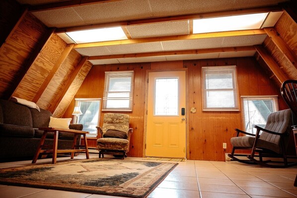 A cozy attic room with a sloped ceiling, wooden walls, a sofa, two rocking chairs, and a door with a window. - Lazy Lane Cabins - Pleasant View (Logan)