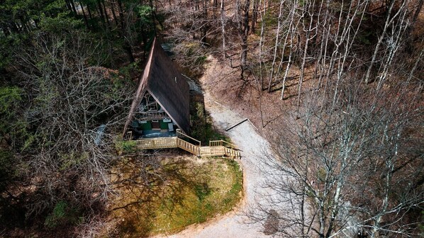 Une vue aérienne d’un chalet doté d’une terrasse en bois et d’un sentier de terre menant à celui-ci, le tout entouré d’une forêt dense.