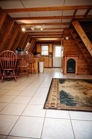 A cozy attic-style room with a fireplace, wooden beams, a tiled floor, and a dining area with chairs. - Lazy Lane Cabins - Pleasant View (Logan)