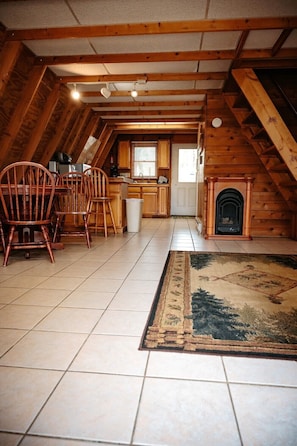 Une pièce confortable, aménagée dans les combles, avec une cheminée, des poutres apparentes, un sol carrelé et un coin repas avec des chaises.