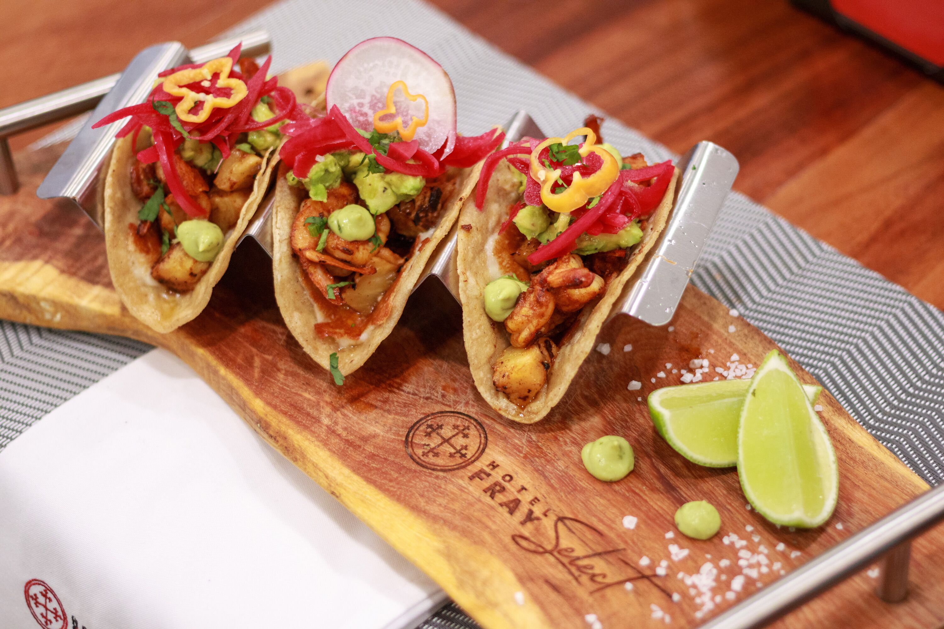 Trois tacos présentés sur une planche en bois, accompagnés d’un logo, de quartiers de citron vert et d’une serviette.
