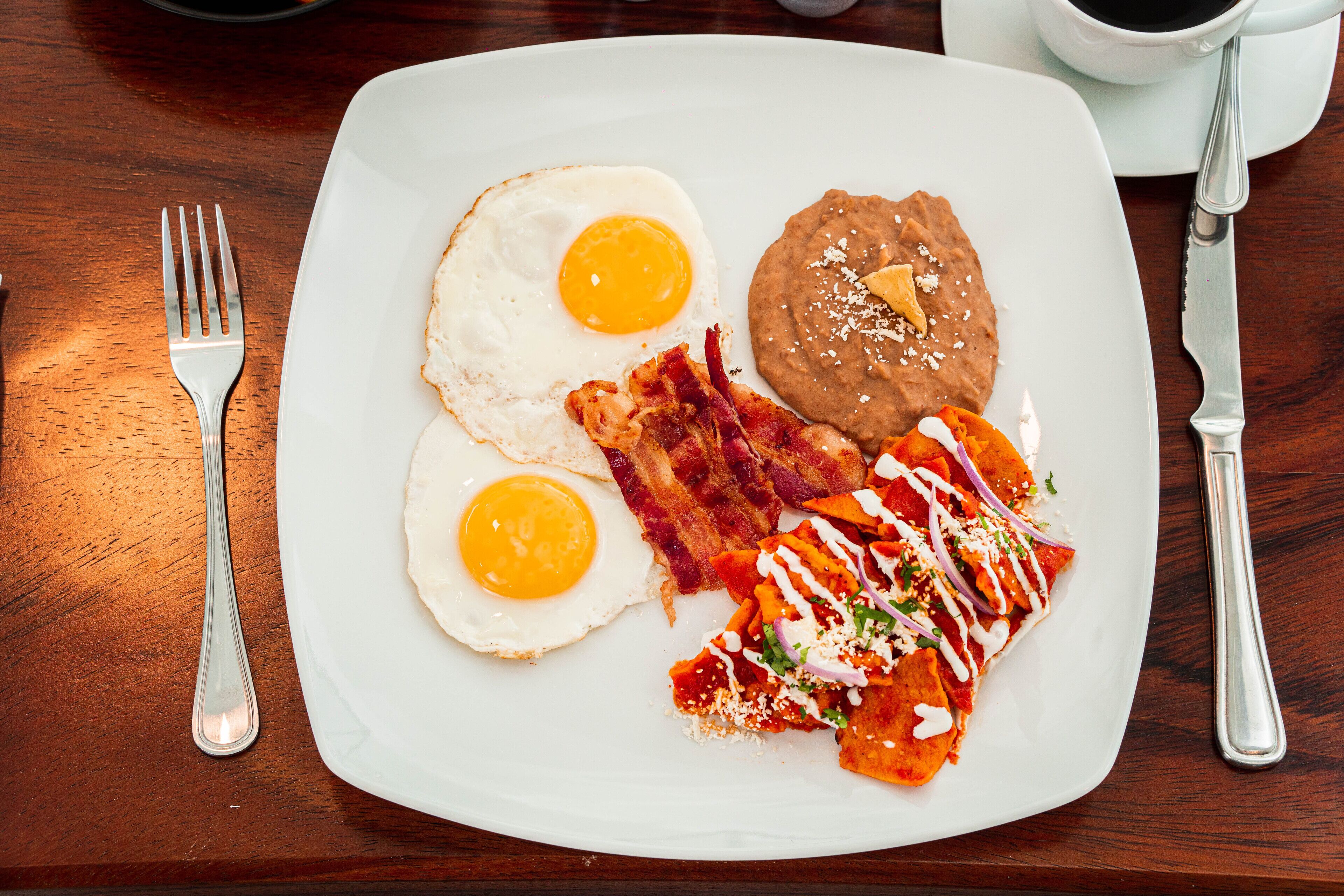 Ein weißer Teller mit zwei Spiegeleiern, Speck, einer Portion Bohnen und einer Schale Chilaquiles mit roter Soße und Käse.
