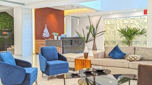 Een moderne lobby met blauwe fauteuils, een bank en een salontafel. In de achtergrond staan planten en een decoratieve boom.