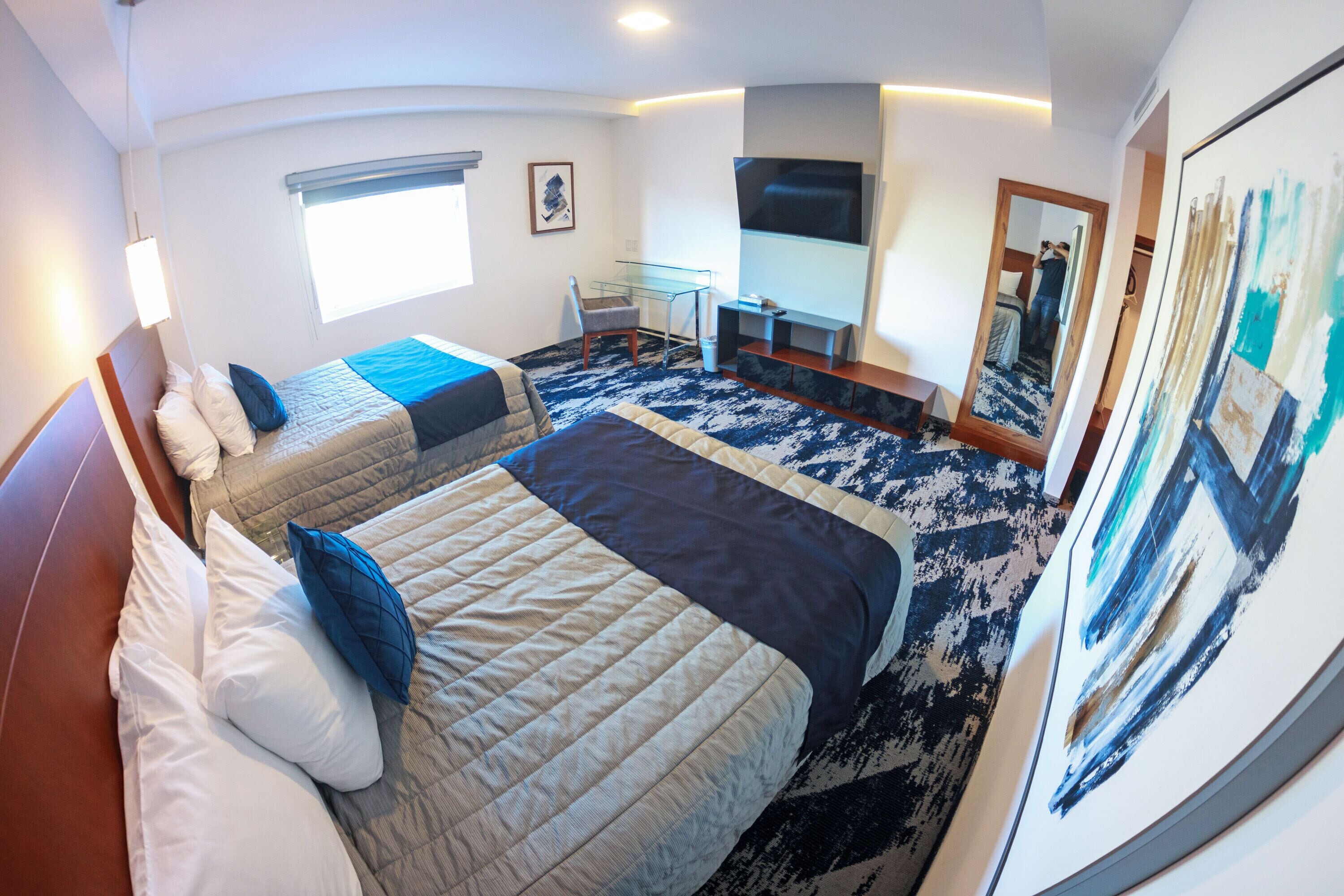 Habitación de hotel con dos camas, un escritorio, una silla, un televisor y un espejo grande.