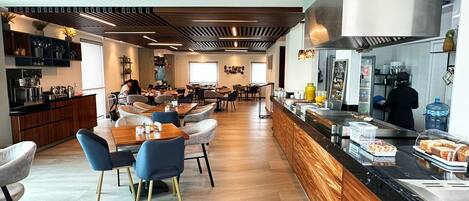 A modern dining area with wooden tables and chairs, a kitchen counter with food items, and a bar area.