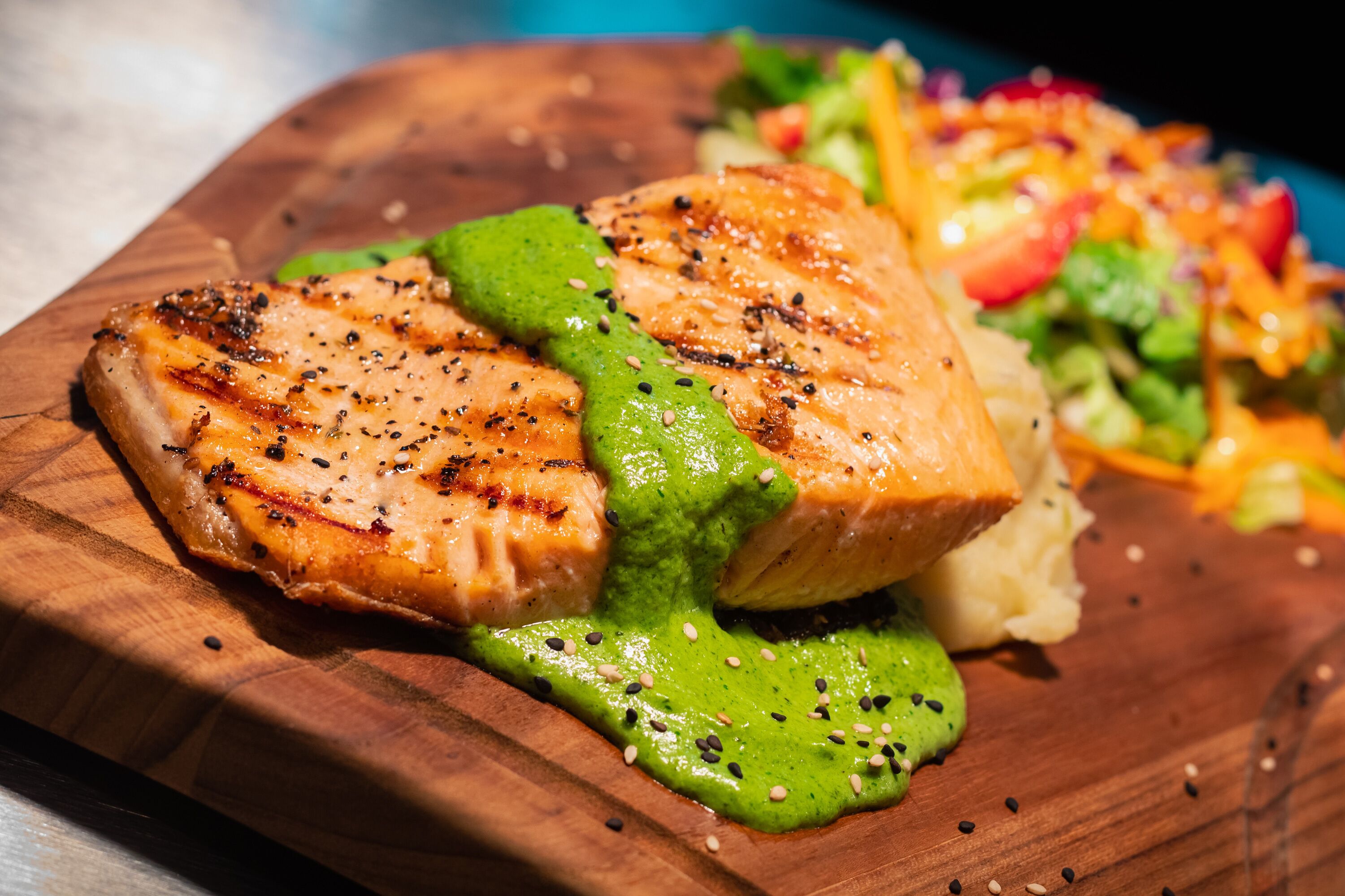 Grilled salmon with green sauce on a wooden board, accompanied by a colorful salad.